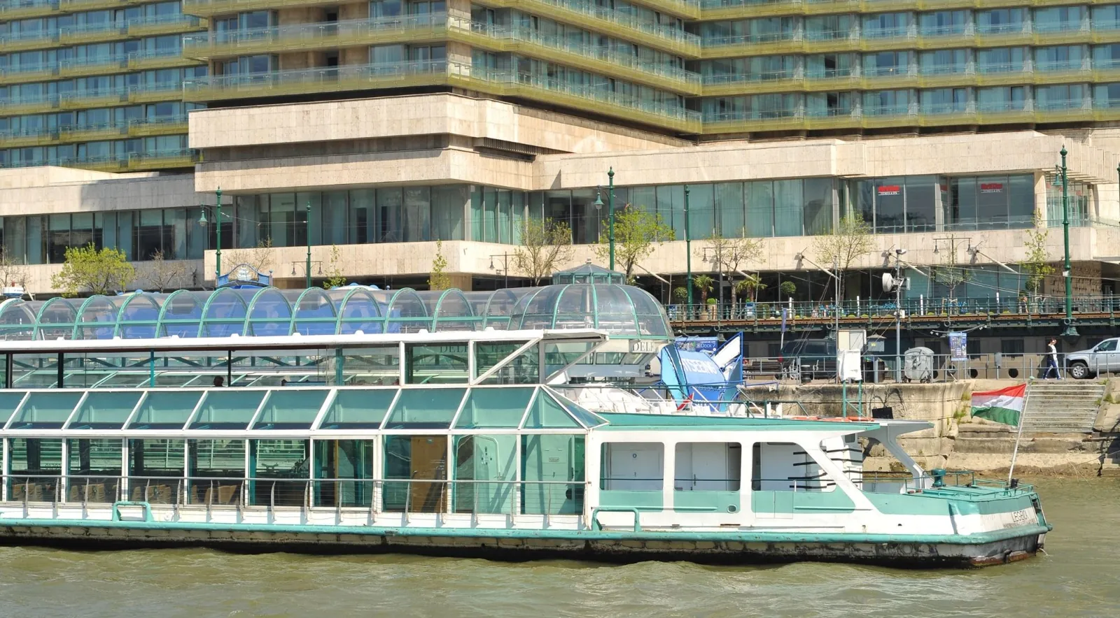 A glass-roofed cruise boat flying the Hungarian flag at a Pest-side pier